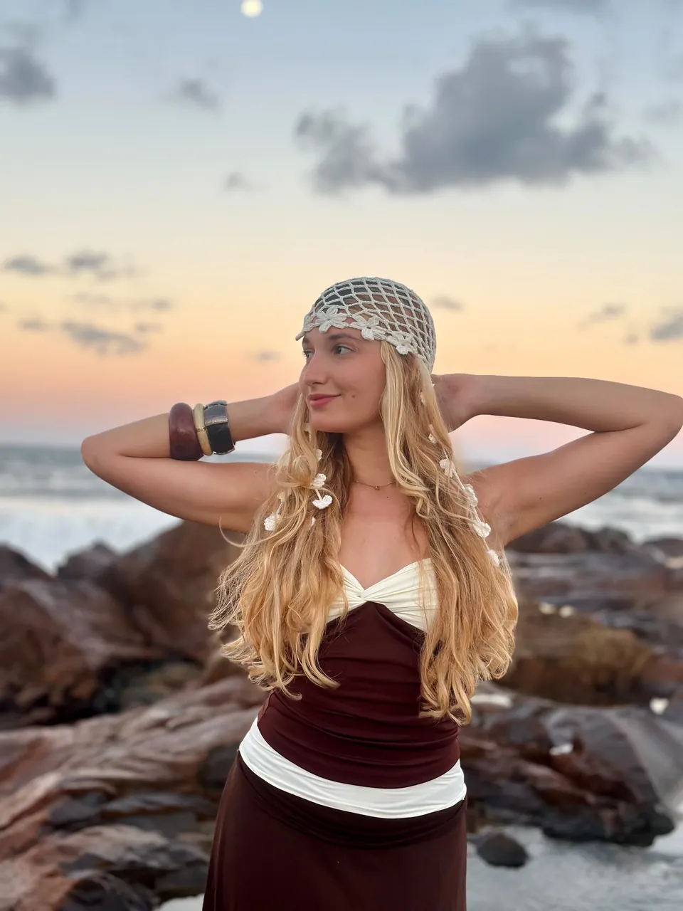 Clara ao pôr do sol nas pedras da praia, com uma rede de crochê no cabelo
